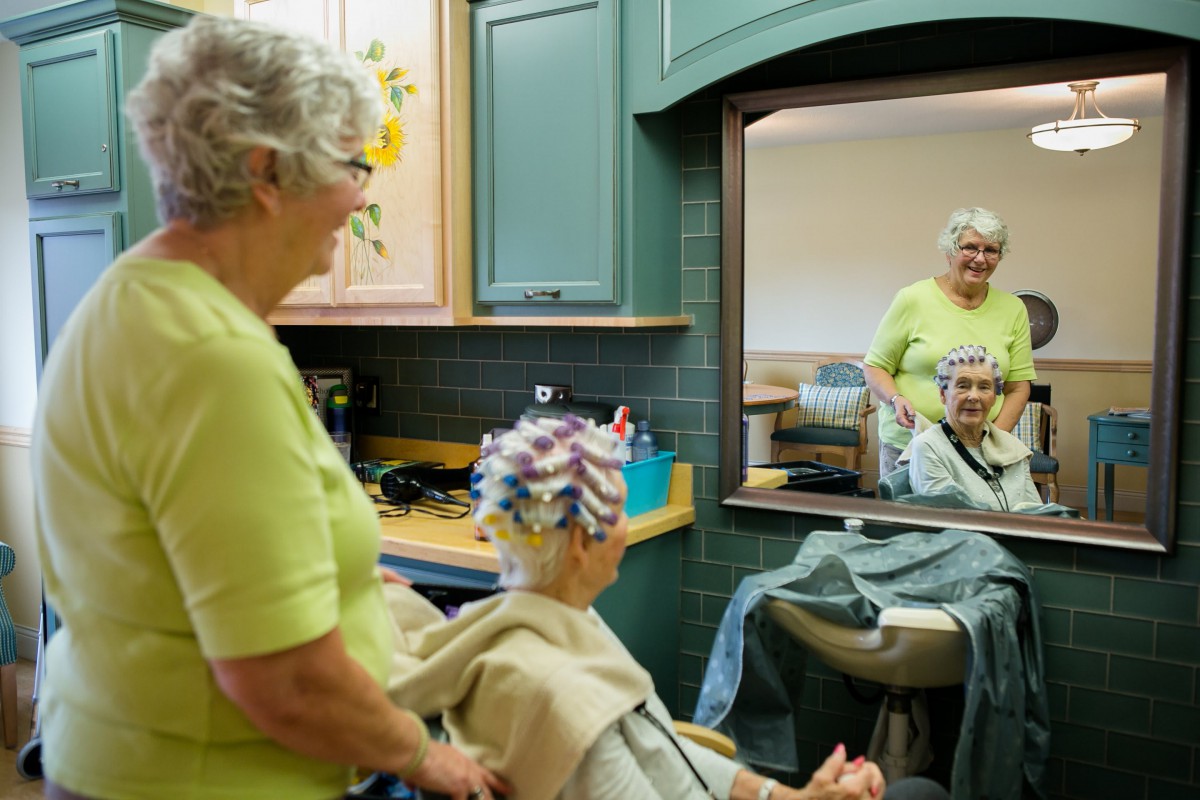 An elderly woman getting her hair styled in a salon, smiling at her reflection in the mirror.
