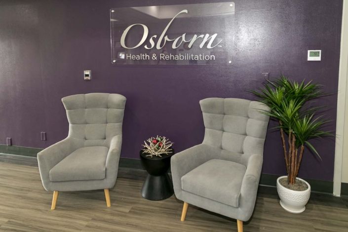 A cozy waiting area with two grey chairs, a small table with a succulent, and a potted plant against a purple wall.