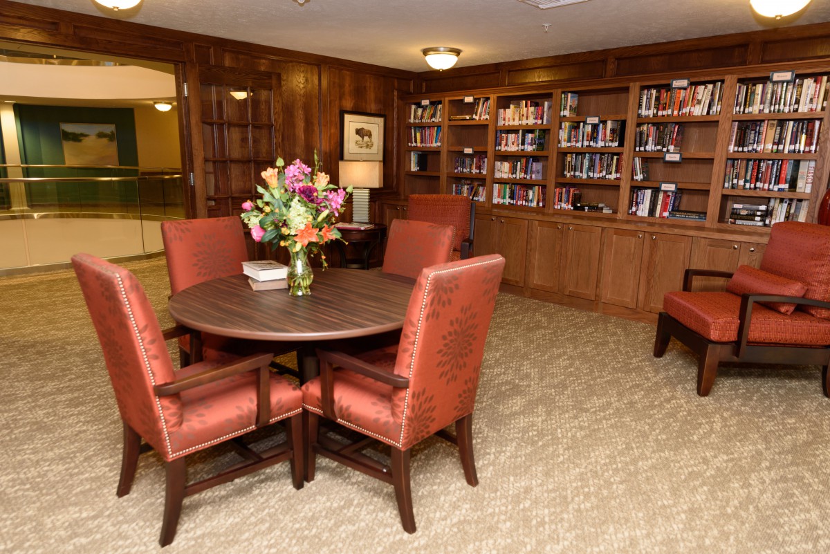 A cozy library with wooden bookshelves, a round table surrounded by upholstered chairs, and a vase of flowers on the table.