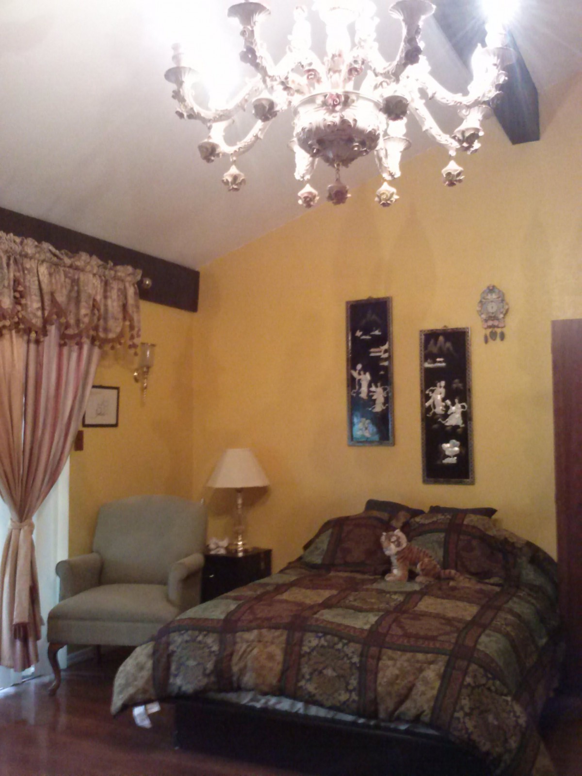 A cozy bedroom with warm yellow walls, a decorative chandelier, and an upholstered armchair.
