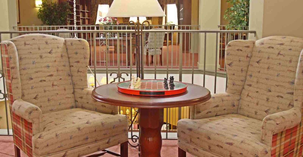 A cozy indoor setup featuring a chess and checkers board on a round wooden table between two upholstered chairs.