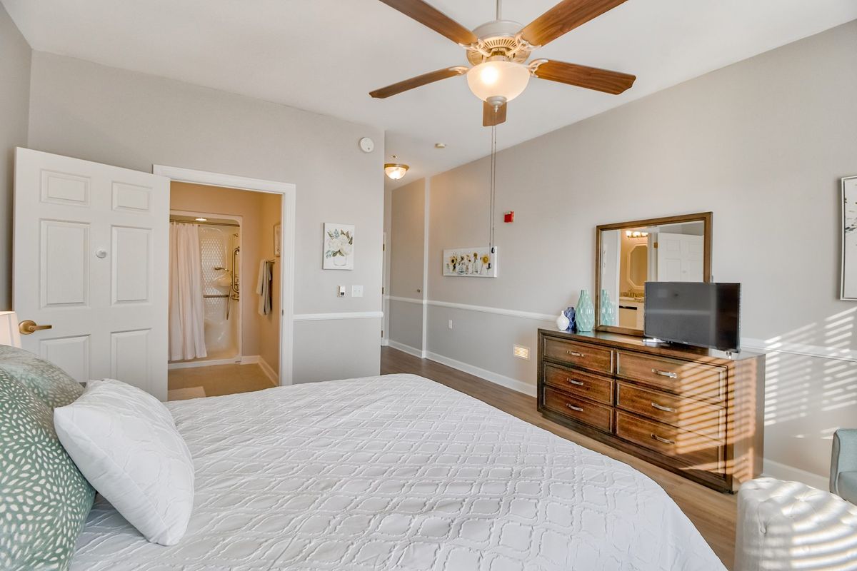 A modern bedroom featuring a wooden dresser, mirror, ceiling fan, and white bedding.