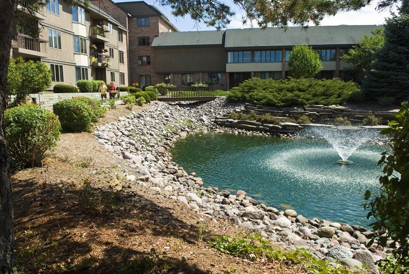 A tranquil scene with an apartment building overlooking a small lake with a fountain and rocky shoreline.