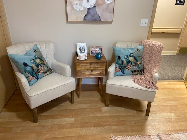 A cozy living room corner featuring two white chairs with decorative pillows, a wooden side table, and wall art.