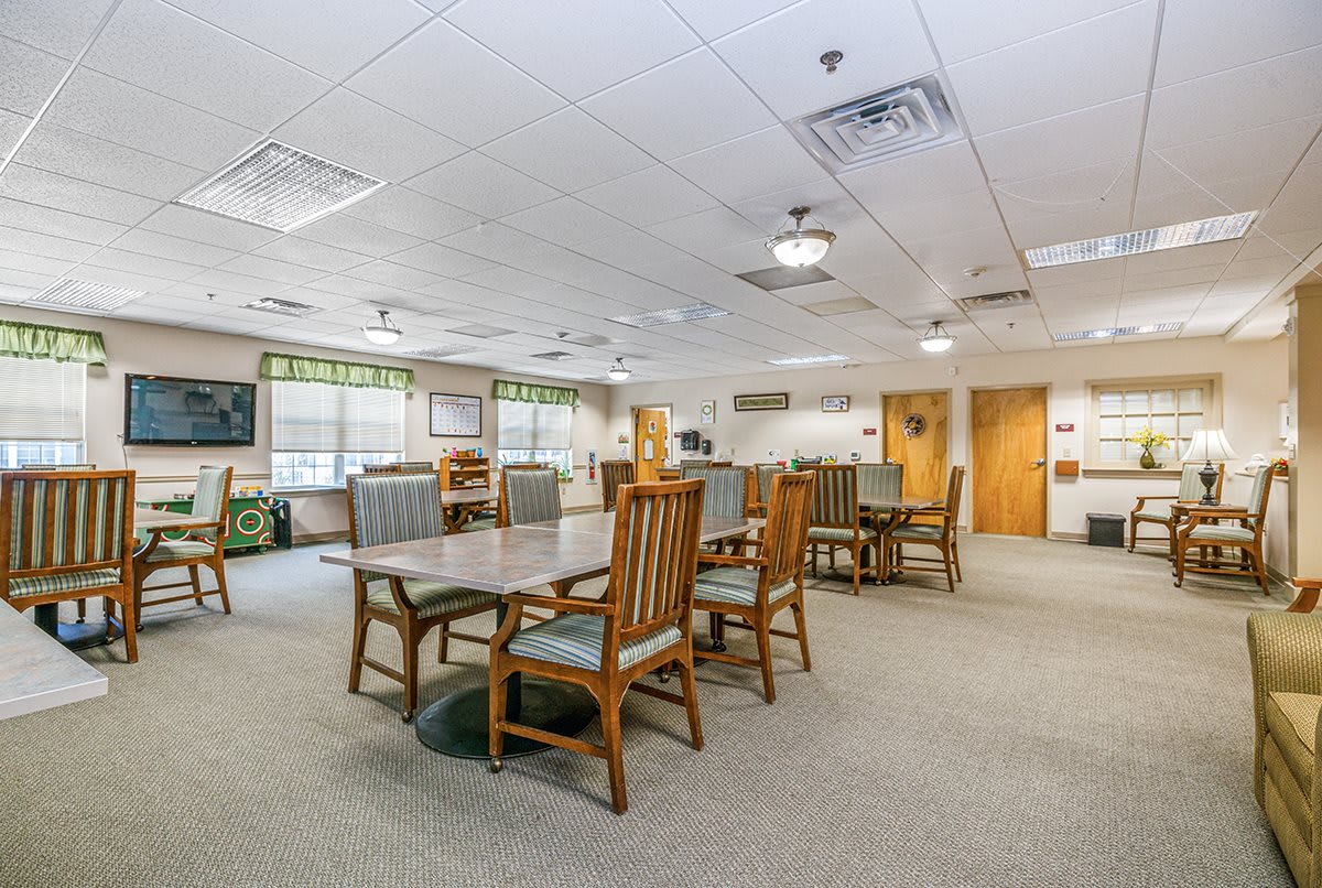 A spacious community room with wooden tables and chairs, featuring large windows and overhead lighting.