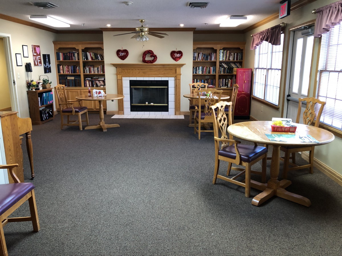 A cozy library reading room with tables, chairs, and a fireplace surrounded by bookshelves.