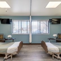A hospital room with two beds separated by a curtain, featuring televisions and light blue walls.