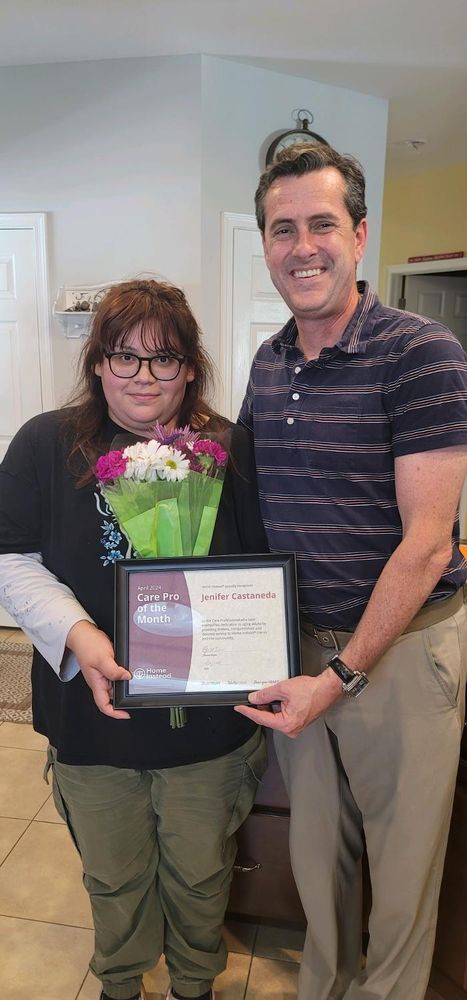 Two people standing together; one is holding a bouquet of flowers and a framed certificate.