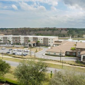 Aerial view of a modern building complex with a parking lot and surrounding trees under a cloudy sky.