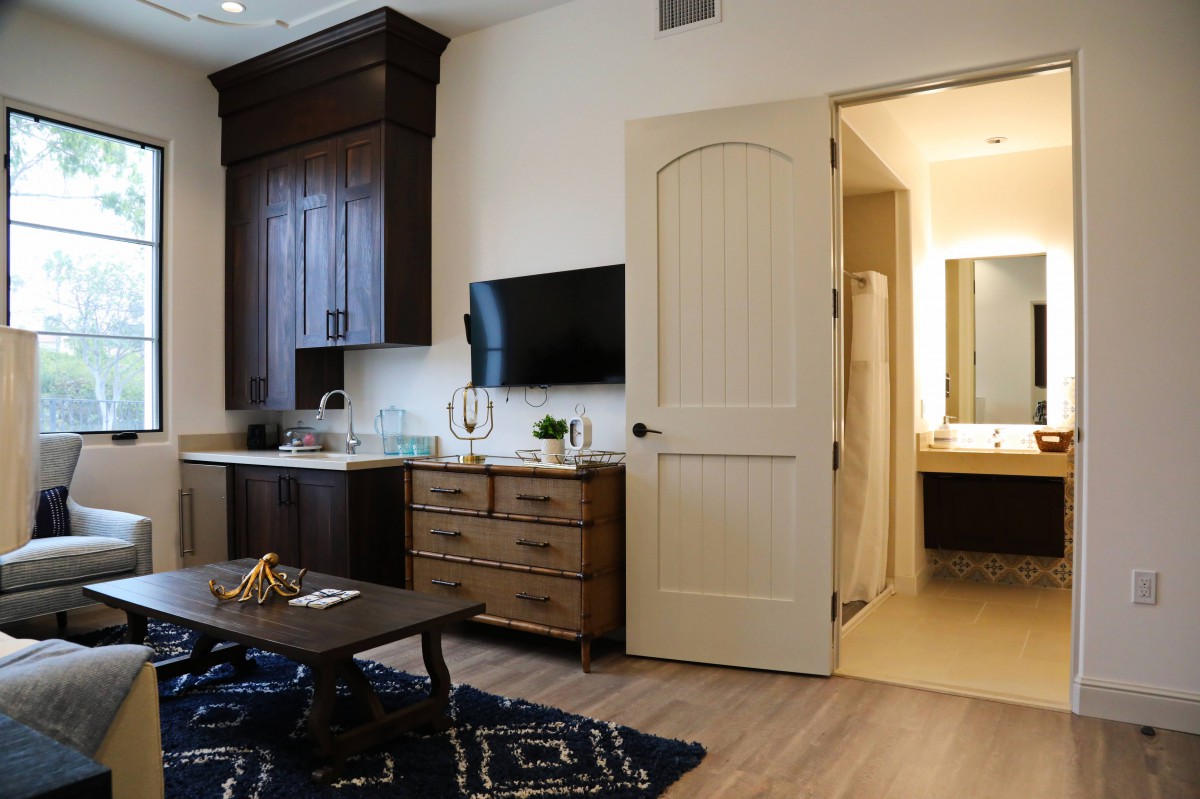 A modern living space featuring a wall-mounted TV, wooden furniture, and a kitchenette leading to a bathroom.