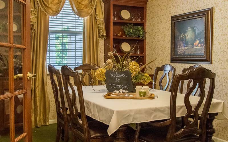 A cozy dining room with a table set for a meal, adorned with a flower centerpiece and surrounded by wooden chairs.
