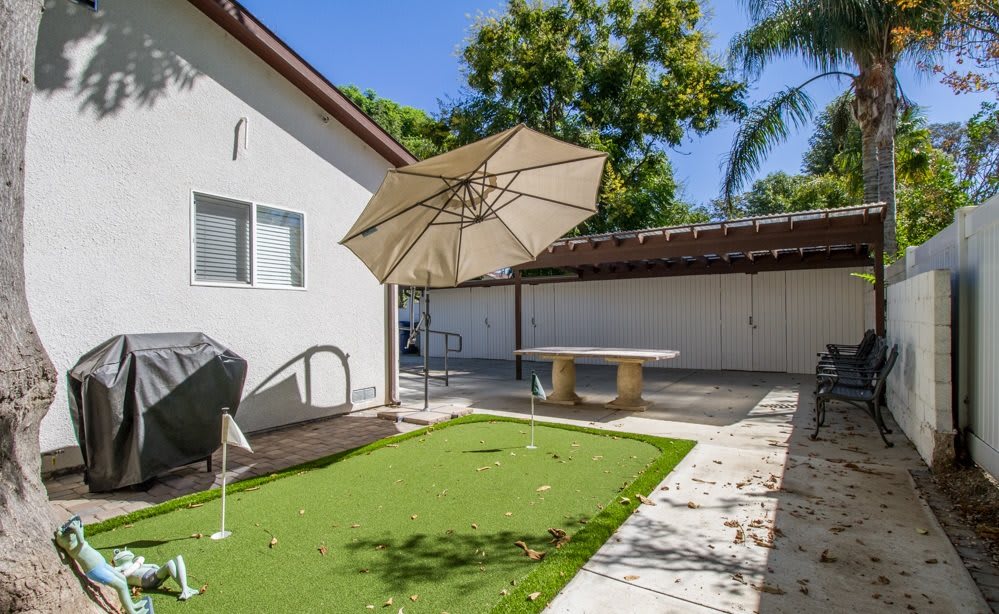 A sunny backyard featuring a small putting green, patio umbrella, and a ping pong table.