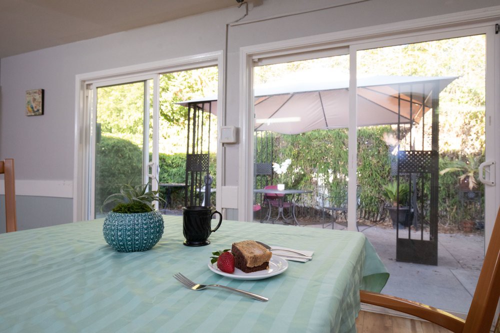 A table set with cake and a strawberry next to a coffee mug, overlooking a patio through large windows.