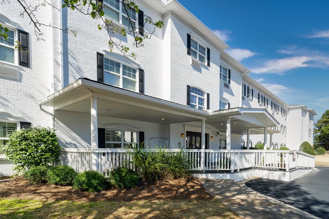A large white residential building with a porch and landscaped front area under a clear blue sky.