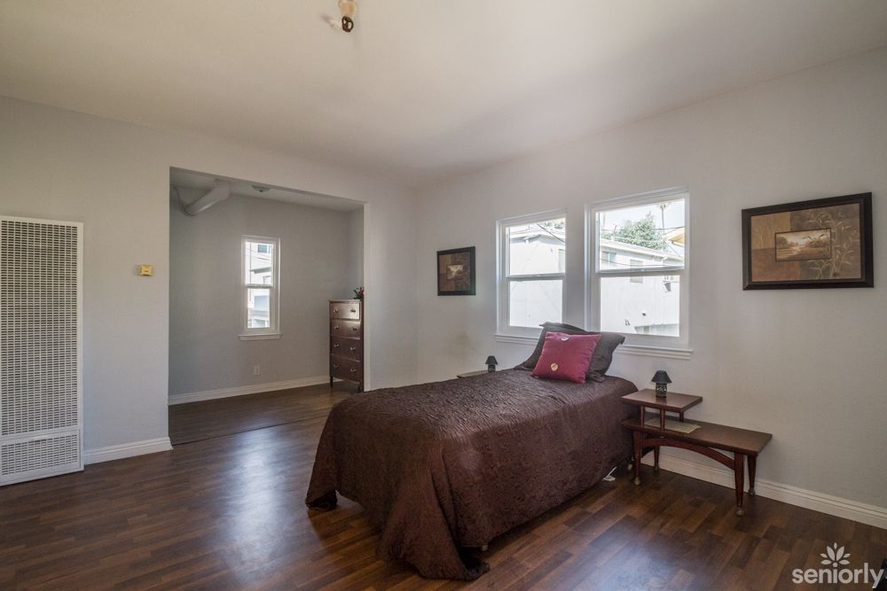 A minimalist bedroom with a single bed, hardwood flooring, and bright natural light coming through the windows.