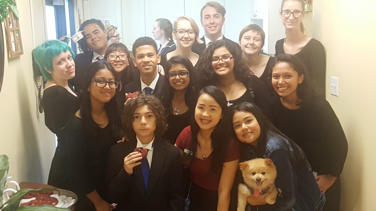 A group of students dressed in formal attire, smiling and posing together indoors with a small dog.