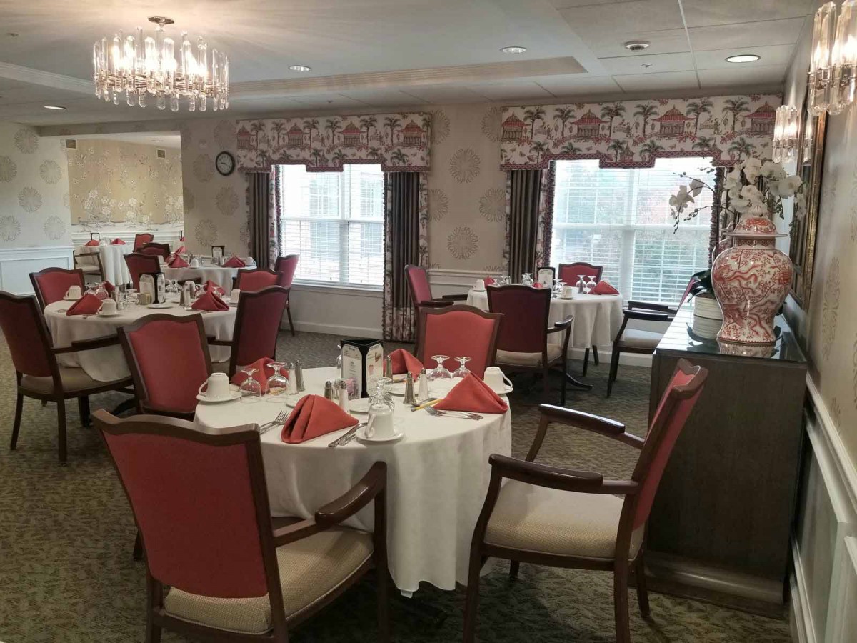 An elegant dining room with tables set for a meal, featuring a chandelier and large windows.