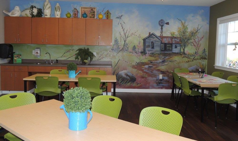 A classroom with a mural depicting a countryside scene, featuring tables and green chairs, complemented by decorative plants.