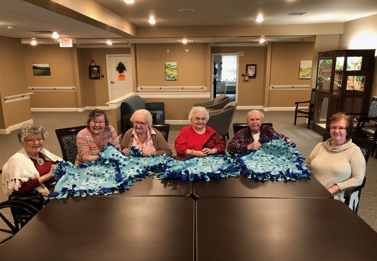 A group of seniors proudly displaying their handcrafted blankets in an indoor setting.