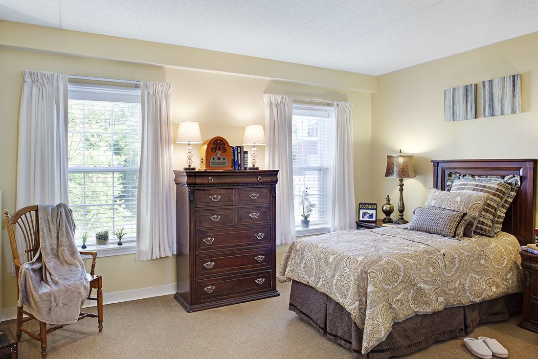 A cozy bedroom with traditional decor featuring a bed, chest of drawers, and two windows with curtains.