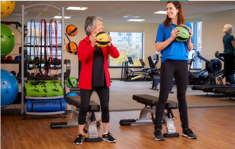 Two women exercising with medicine balls in a gym setting.