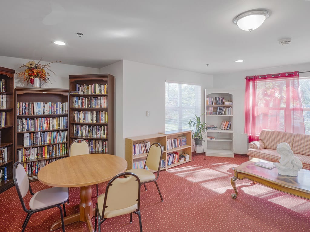 A cozy library reading area with bookshelves, a table, and chairs illuminated by natural sunlight.