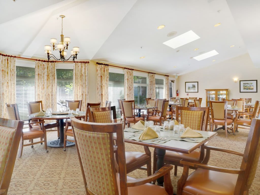 A well-lit dining room with neatly arranged tables and chairs, featuring large windows and a chandelier.