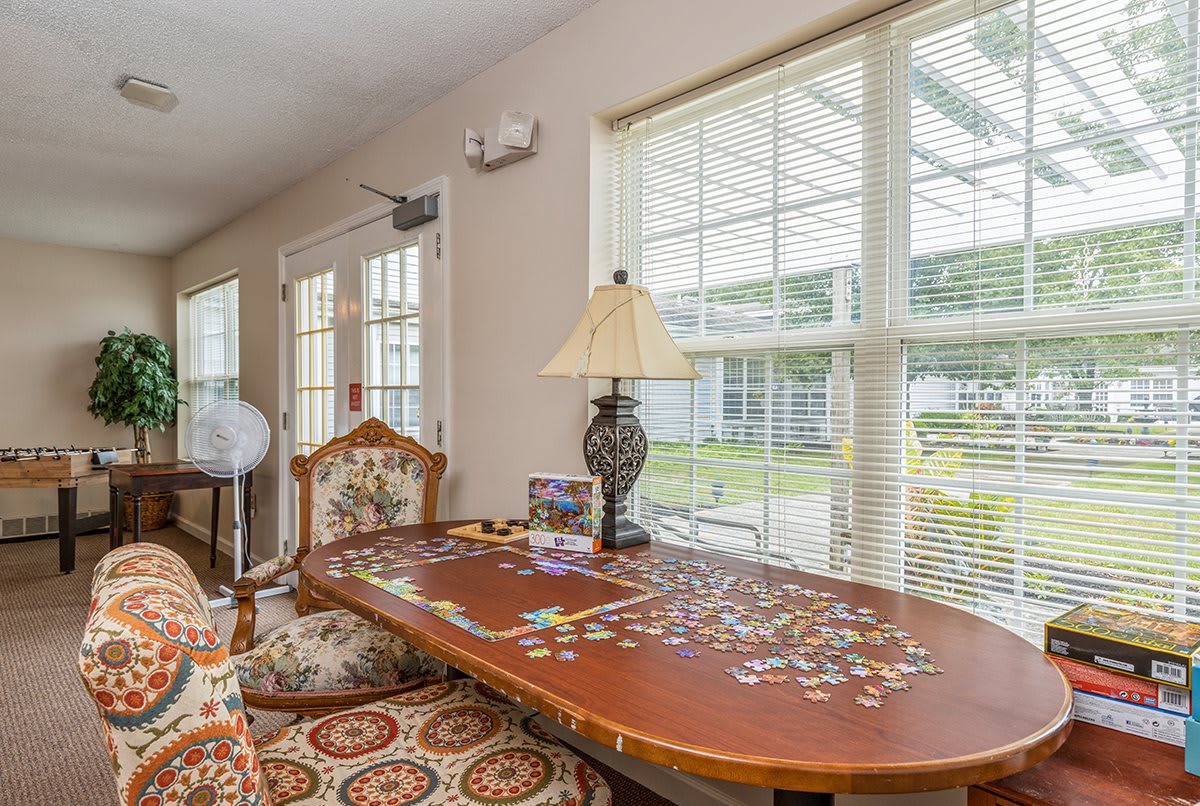 A cozy room with a wooden table covered in a partially completed puzzle, accompanied by an ornate lamp and patterned chair, bathed in natural light from large windows.