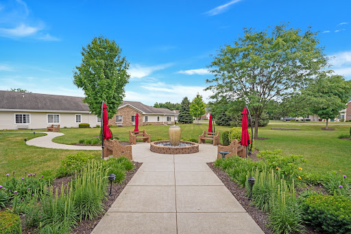A serene garden pathway leading to a fountain, surrounded by lush greenery and vibrant red umbrellas against a clear blue sky.