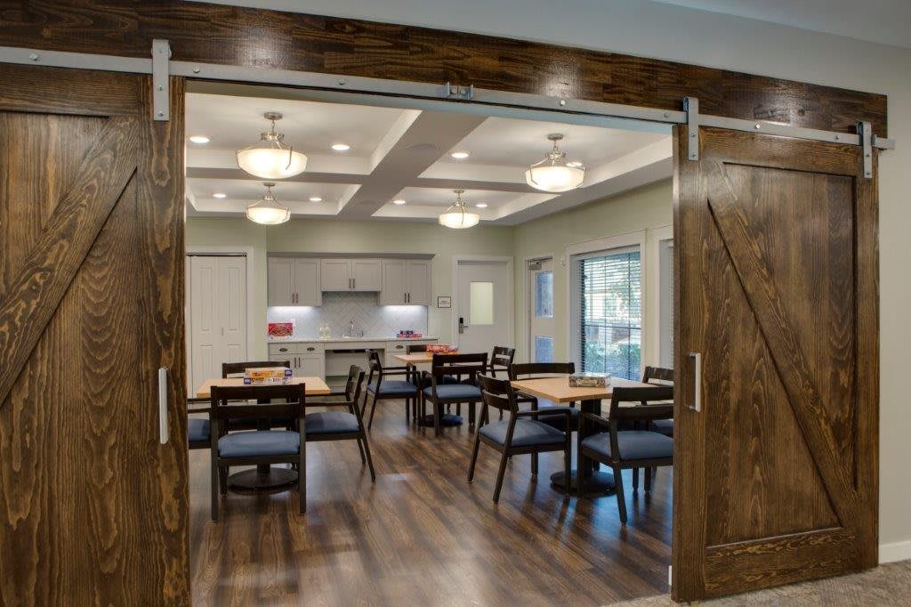 A modern dining area with wooden tables and chairs, connected to a kitchen space, featuring elegant lighting and wood flooring.