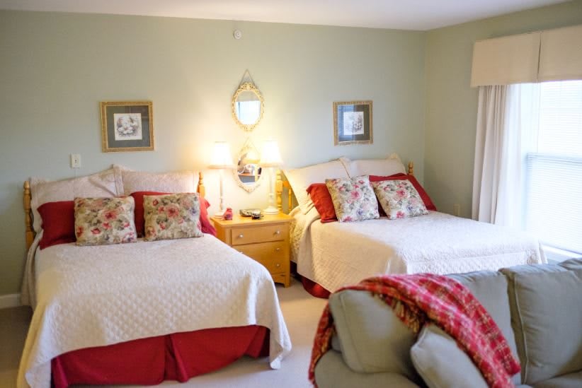 A cozy bedroom featuring two neatly made beds with decorative pillows and a shared nightstand between them, illuminated by warm lamps against a backdrop of soft wall art.