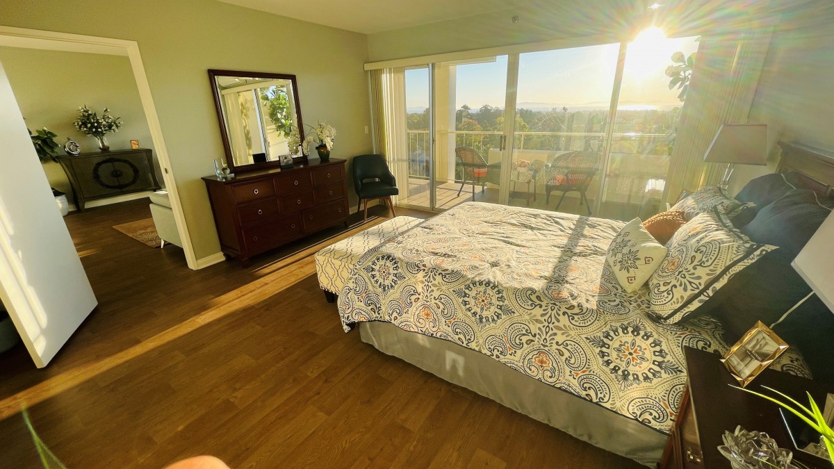 A sunlit bedroom with a large bed, dresser, and a view from the balcony.