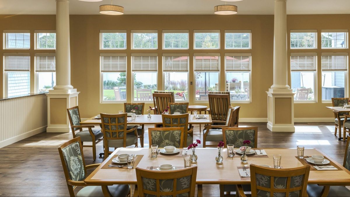 An empty dining room with neatly arranged tables and chairs, illuminated by natural light through large windows.