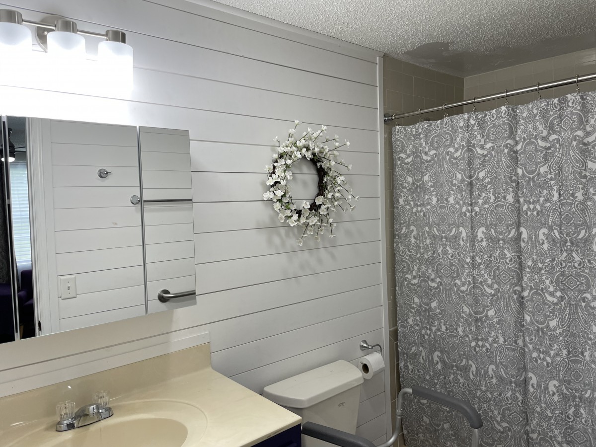 A well-lit bathroom featuring a mirror, decorative wreath on white wood paneling, and a patterned shower curtain.