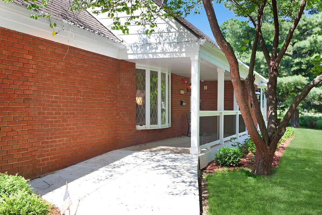 A charming red brick house with a front porch surrounded by trees and landscaping.