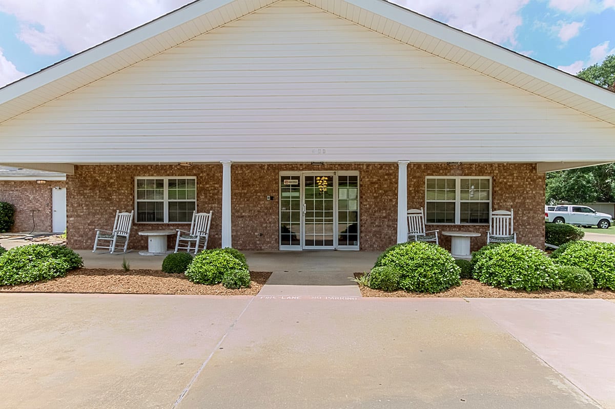 A single-story building with a porch, featuring brick and white siding, two chairs, and neatly trimmed bushes.