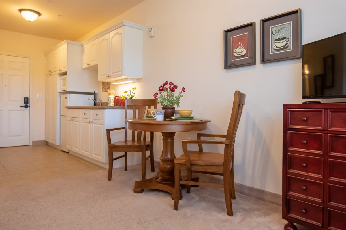 A cozy dining area with wooden furniture adjacent to a kitchen, featuring a small table with chairs and decorative elements like flowers and framed pictures on the wall.