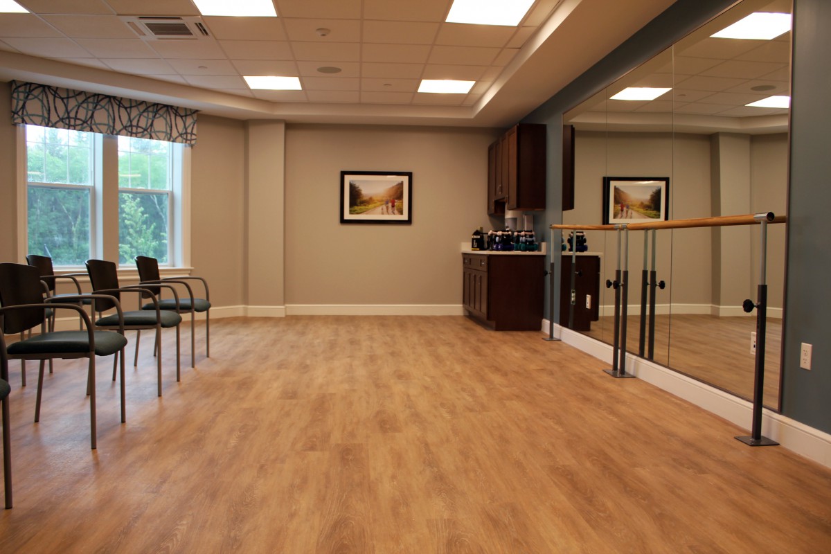 A spacious room with wooden flooring, featuring bright lighting, chairs along the wall, and large mirrors.