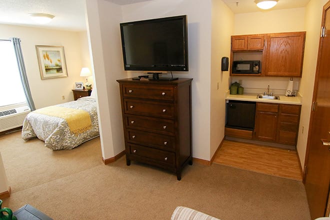 A cozy hotel room featuring a bedroom with a television atop a dresser and a small kitchenette.