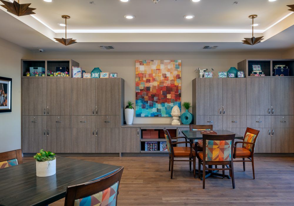 A well-decorated interior space featuring a table and chairs, artwork on the wall, shelves with decor items, and warm lighting.