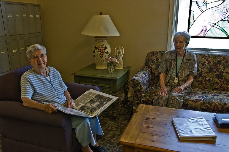 Two elderly women sitting in a cozy living room, one reading a newspaper.