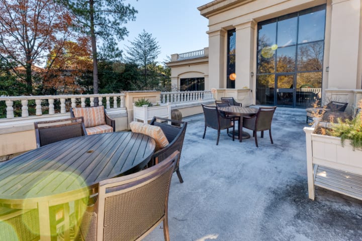 A sunny outdoor patio with tables and chairs, surrounded by elegant architecture and trees.