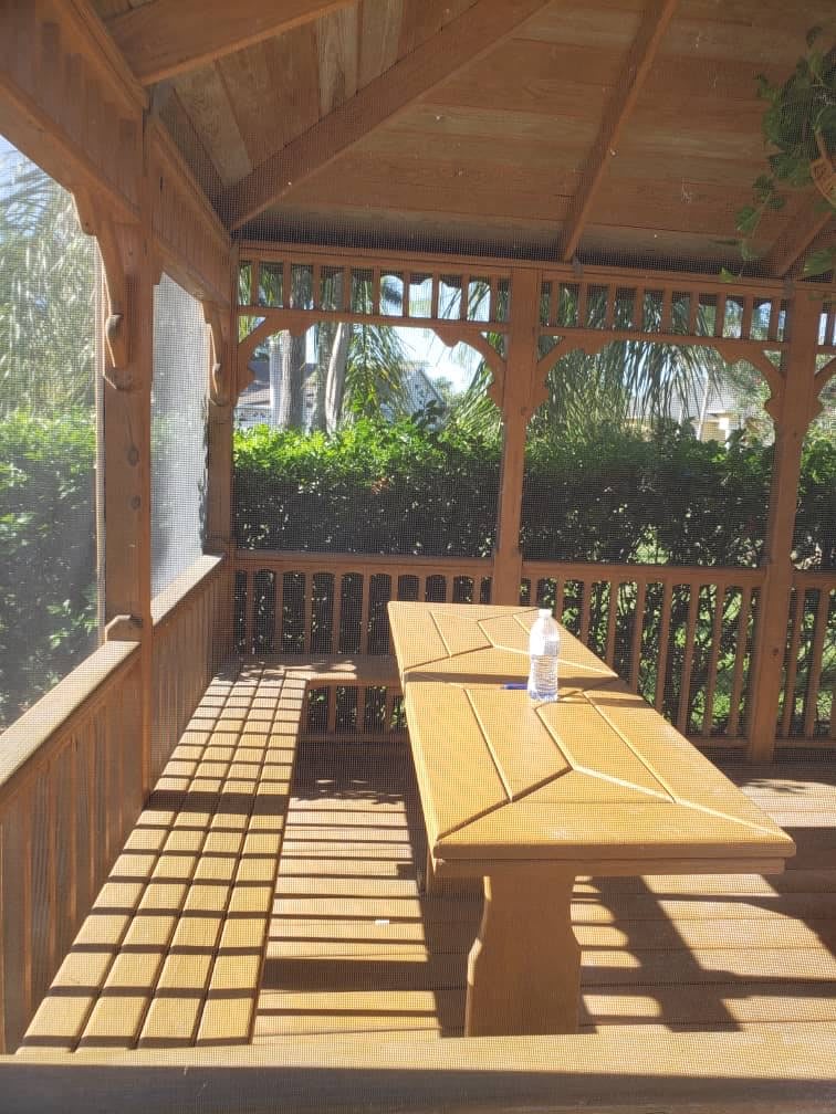 A wooden gazebo with a bench and table, surrounded by greenery, bathed in sunlight.