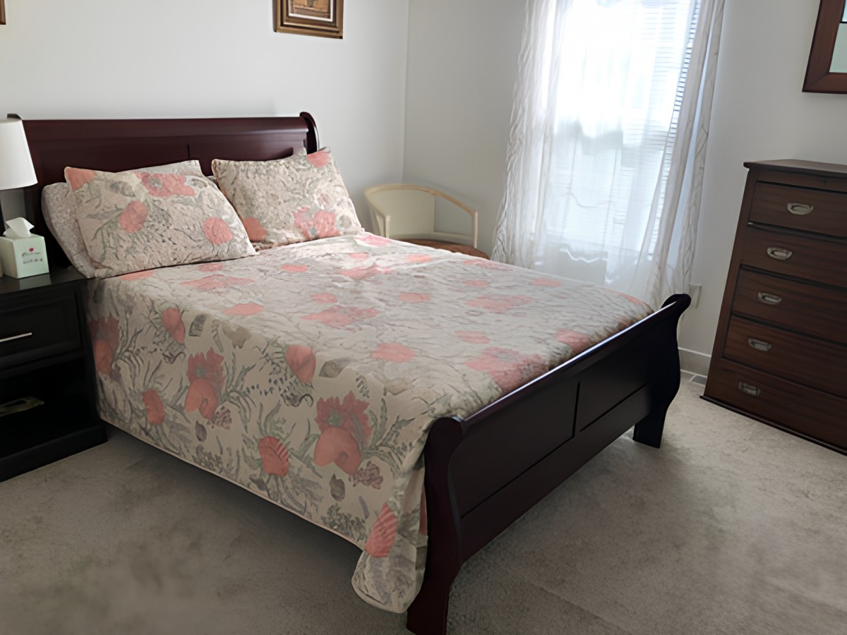 A cozy bedroom featuring a wooden bed with floral bedding and a window with sheer curtains.