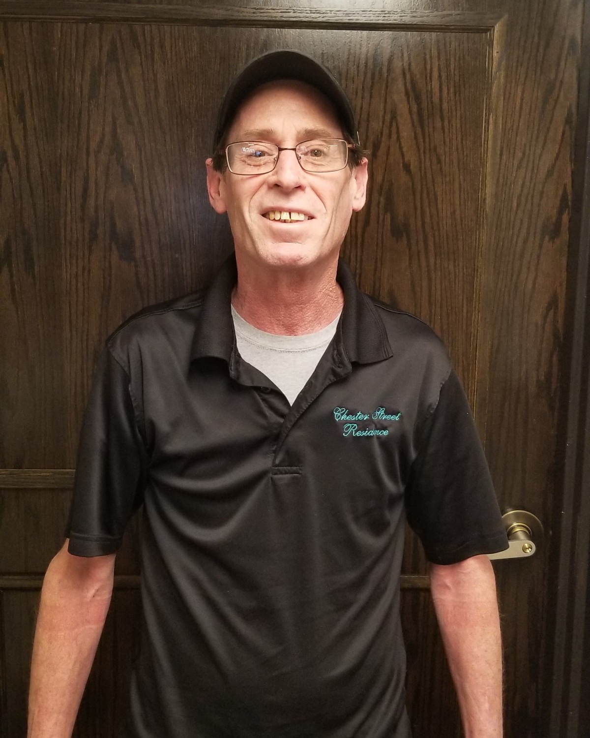 A man wearing a black cap and shirt, smiling in front of a wooden door.