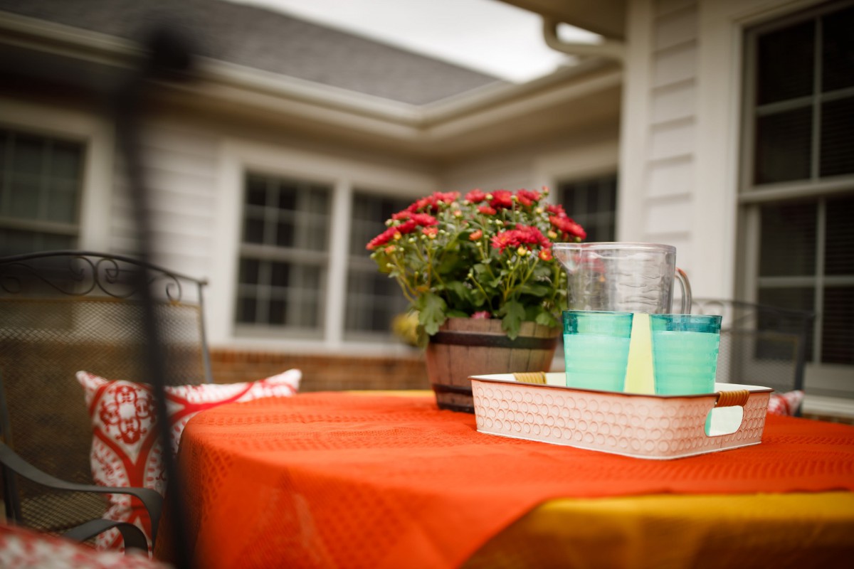 An outdoor table decorated with a vibrant orange tablecloth, featuring a potted plant with red flowers and stacks of books alongside glasses.