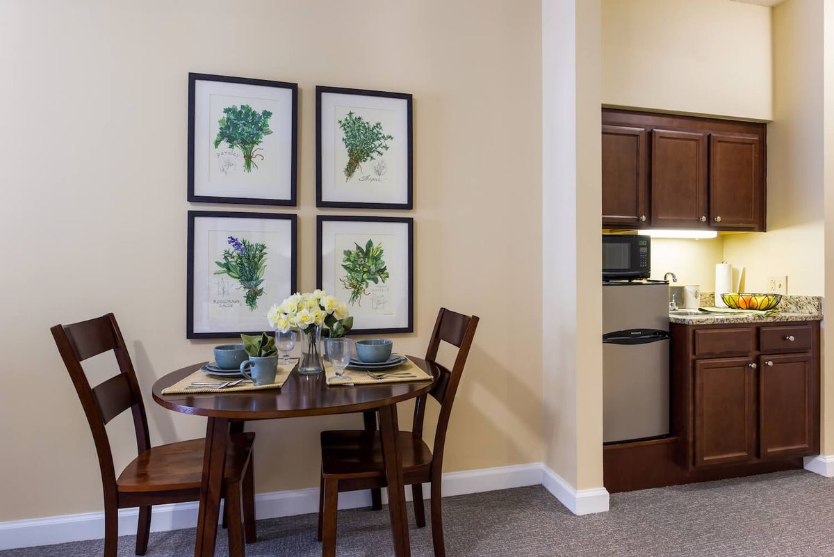 A cozy dining area with a small table set for two, featuring flower decorations and framed botanical art on the wall, adjacent to a kitchenette.