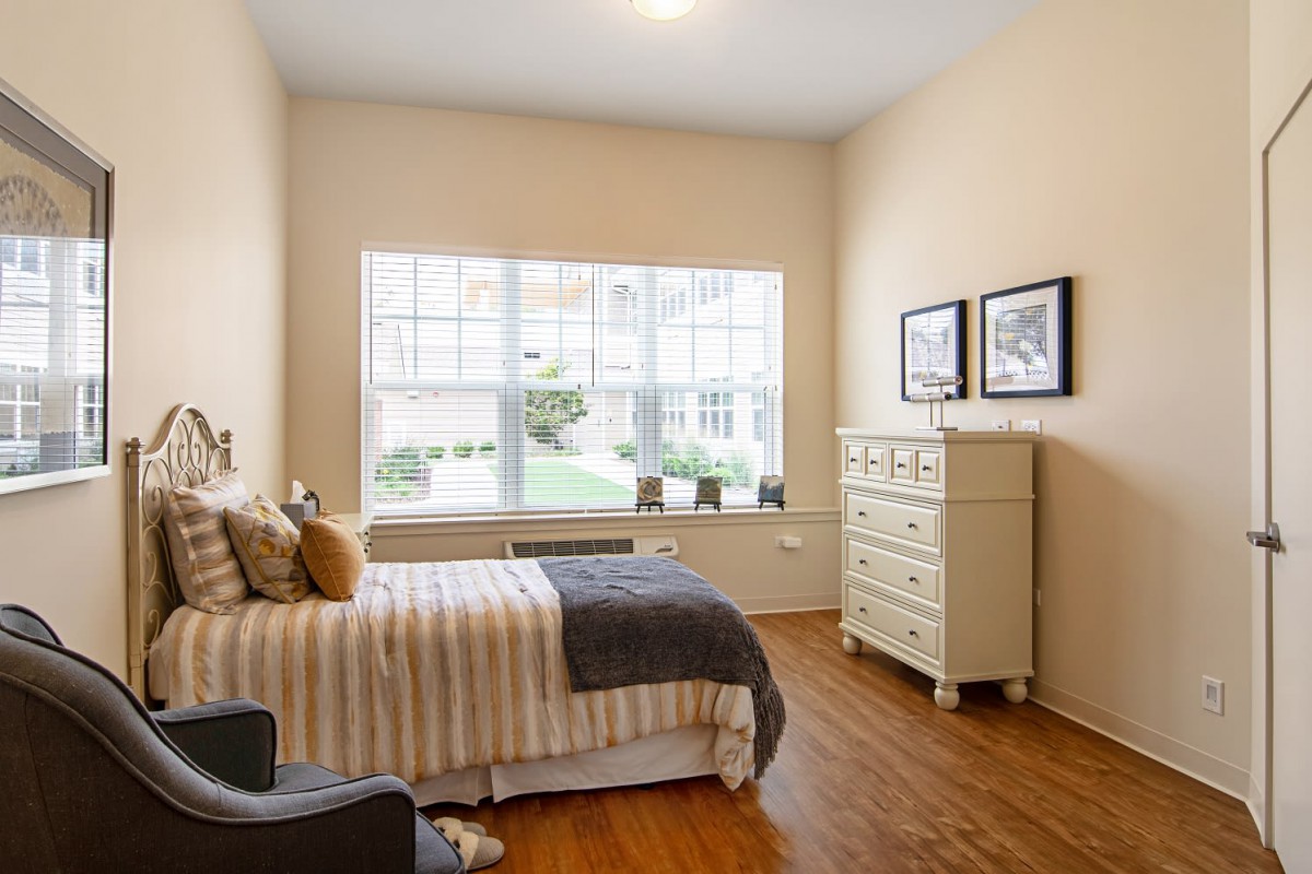 A cozy bedroom with a large window, featuring a bed with striped bedding and a white dresser on a wooden floor.