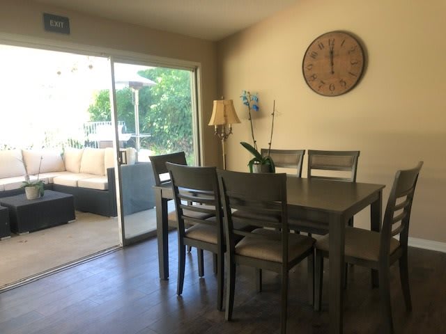 A cozy dining area with a wooden table and chairs, featuring a wall clock and an indoor plant, next to a glass sliding door leading to an outdoor patio.