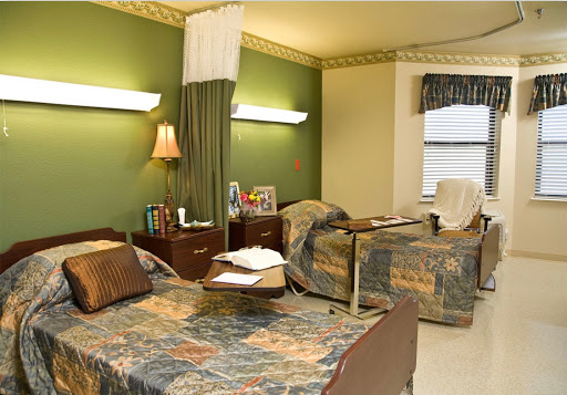 A hospital patient room featuring two beds with decorative covers and a calming green wall.